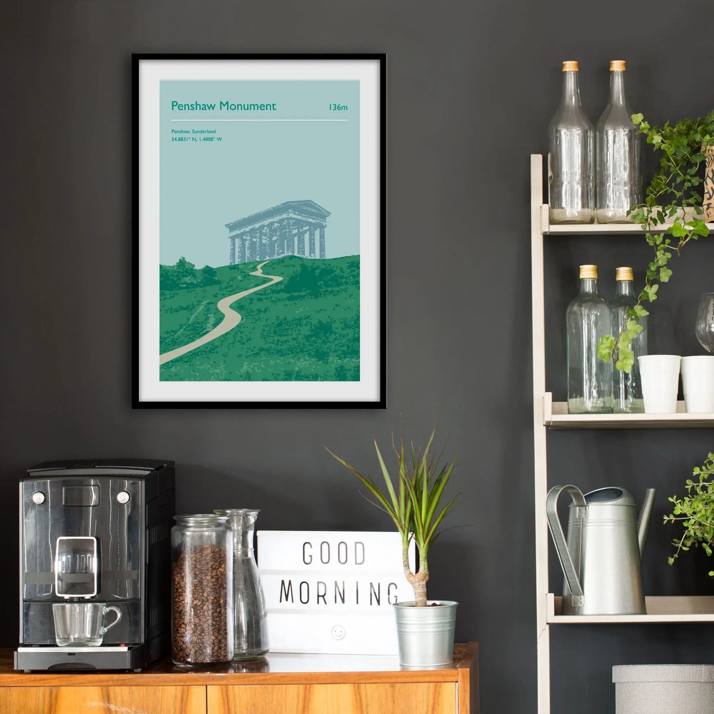 Kitchen wall infront of which is a sideboard with a coffee machine and a set of shelves holding bottles and plants. On the wall above the sideboard is the framed print in a black frame on a dark grey wall.