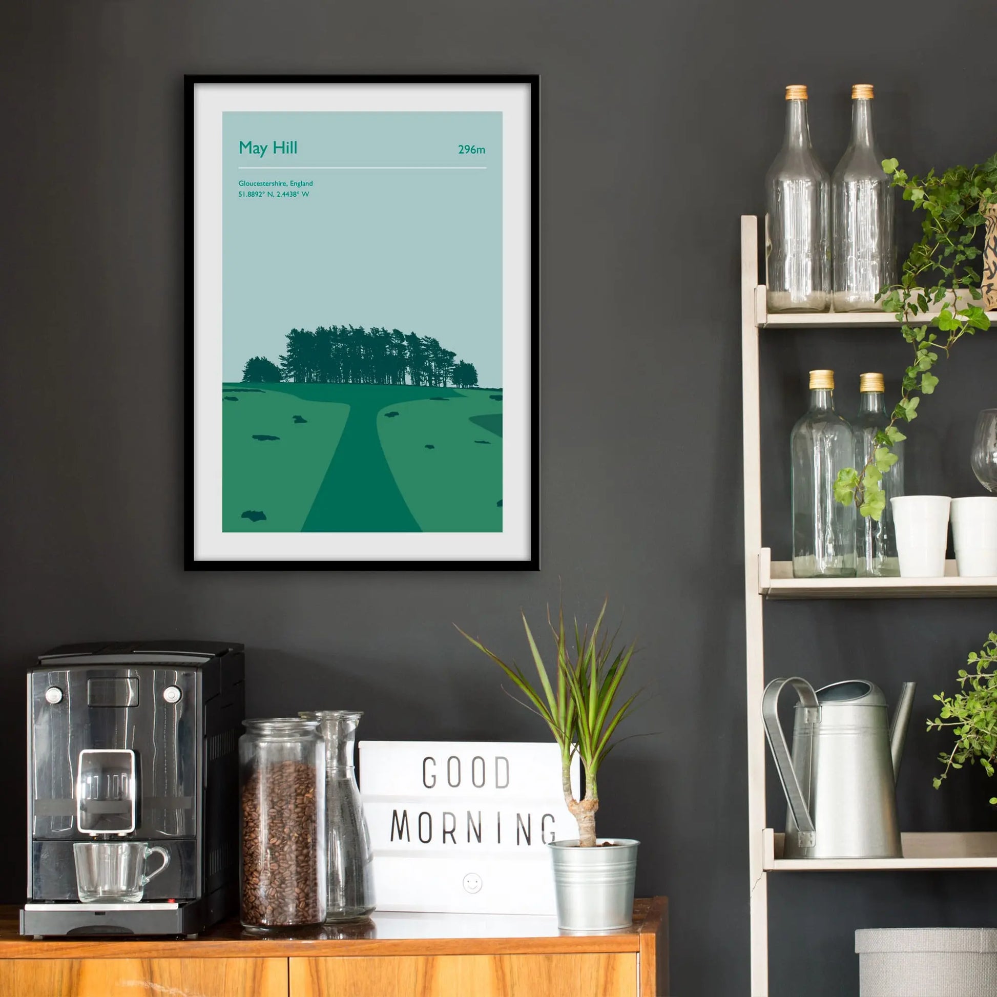 Kitchen wall infront of which is a sideboard with a coffee machine and a set of shelves holding bottles and plants. On the wall above the sideboard is the framed print in a black frame on a dark grey wall.