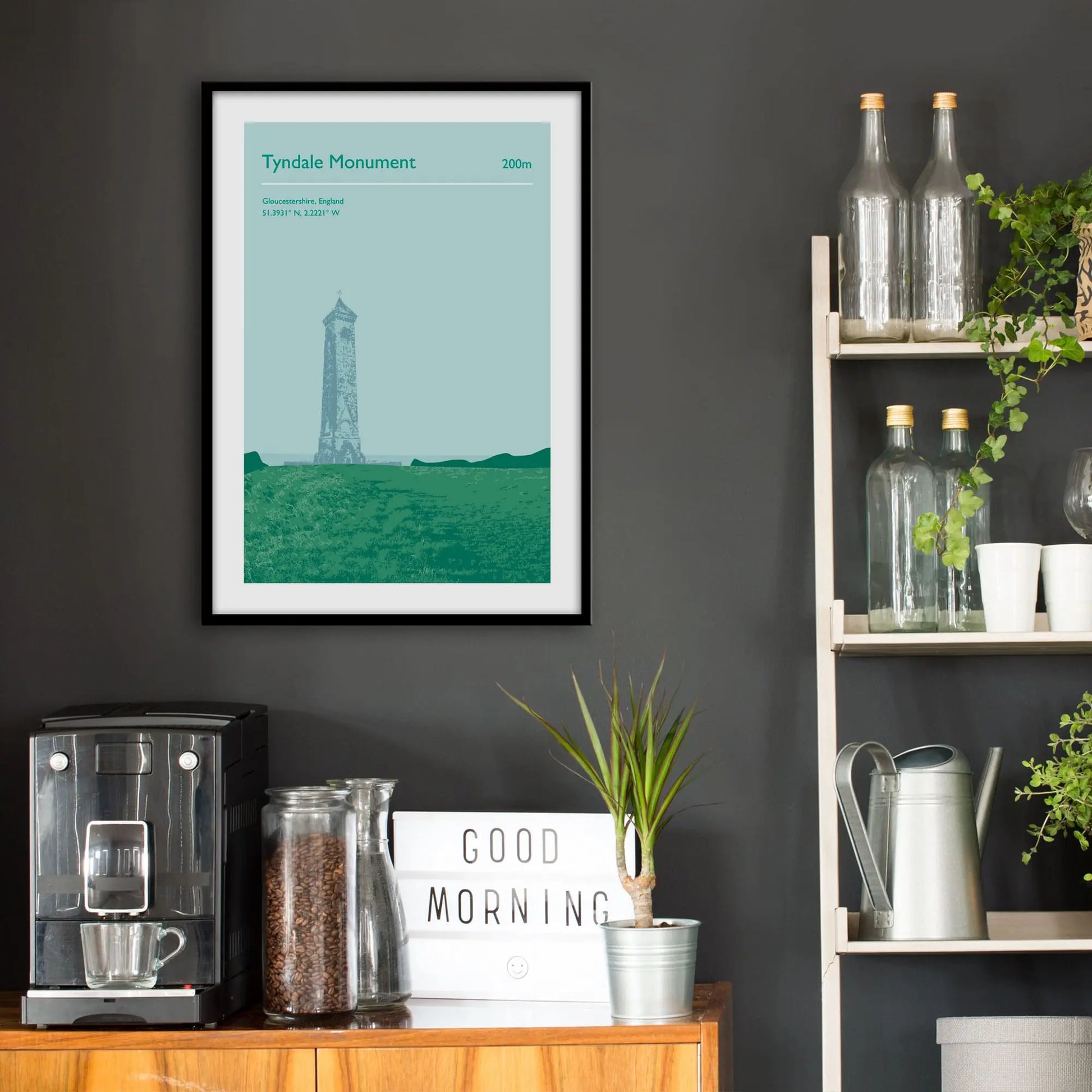 Kitchen wall infront of which is a sideboard with a coffee machine and a set of shelves holding bottles and plants. On the wall above the sideboard is the framed print in a black frame on a dark grey wall.