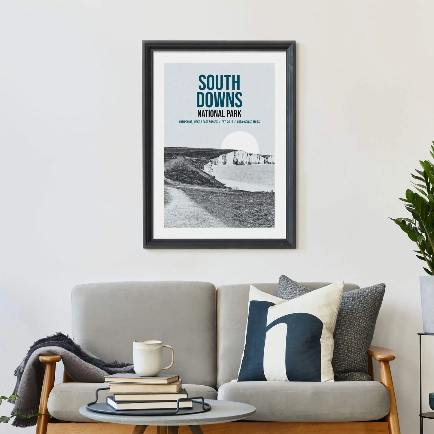 Large print on wall behind a 2 seater sofa covered in cushions with a coffee table covered in books.