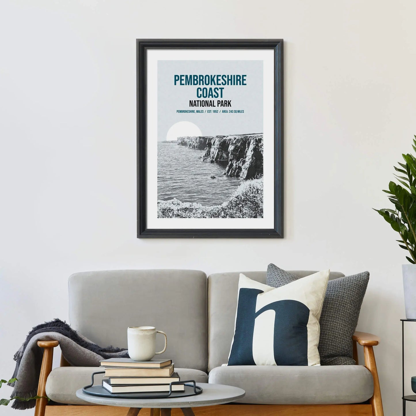 Large print on wall behind a 2 seater sofa covered in cushions with a coffee table covered in books.