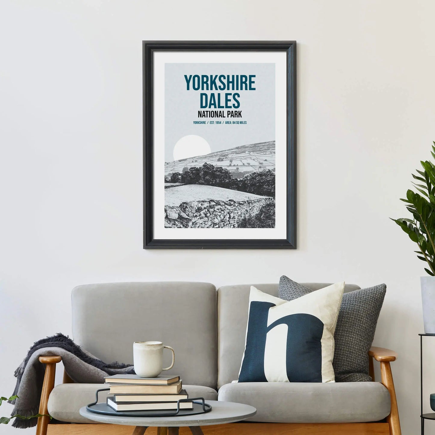 Large print on wall behind a 2 seater sofa covered in cushions with a coffee table covered in books.