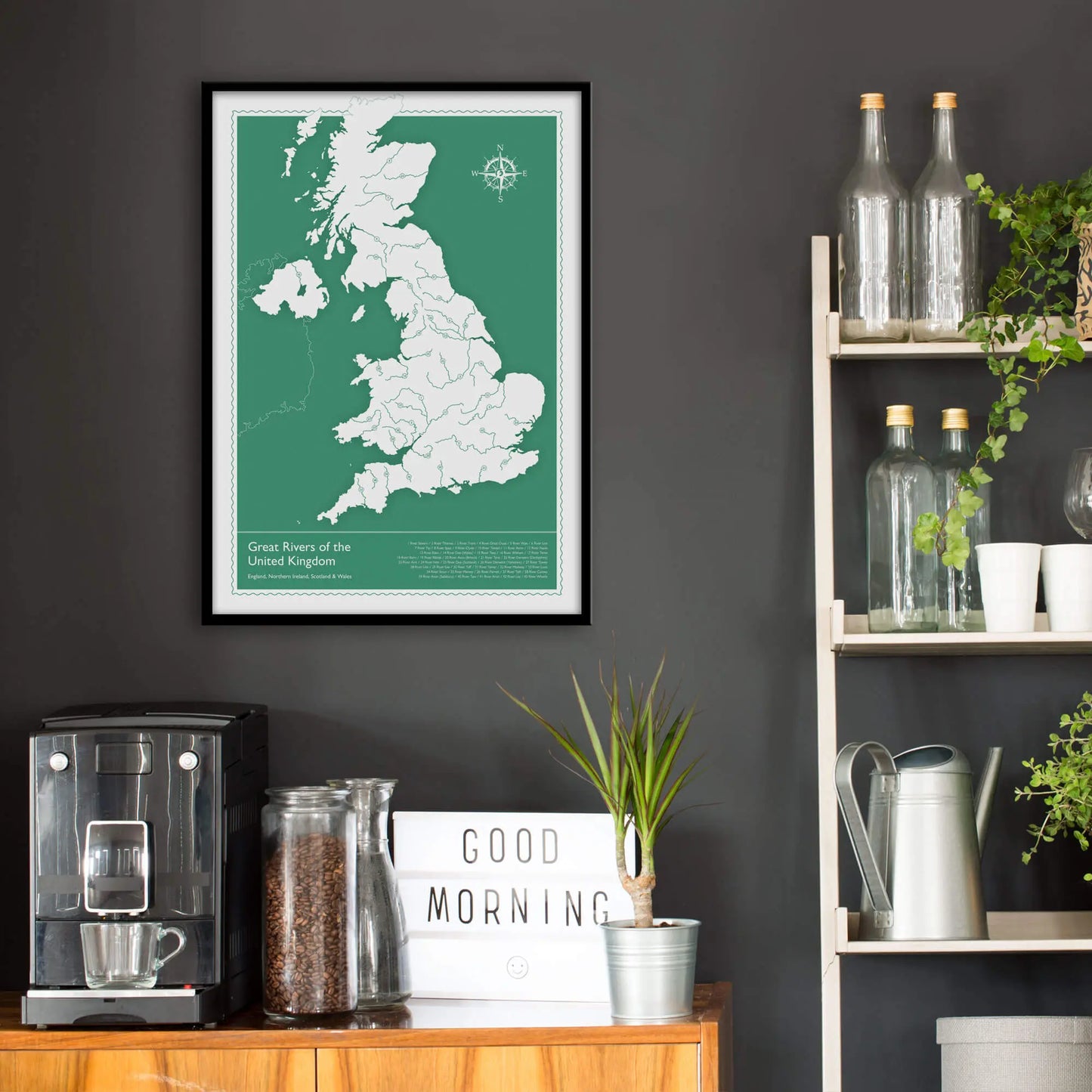 Kitchen wall infront of which is a sideboard with a coffee machine and a set of shelves holding bottles and plants. On the wall above the sideboard is the framed print in a black frame on a dark grey wall.