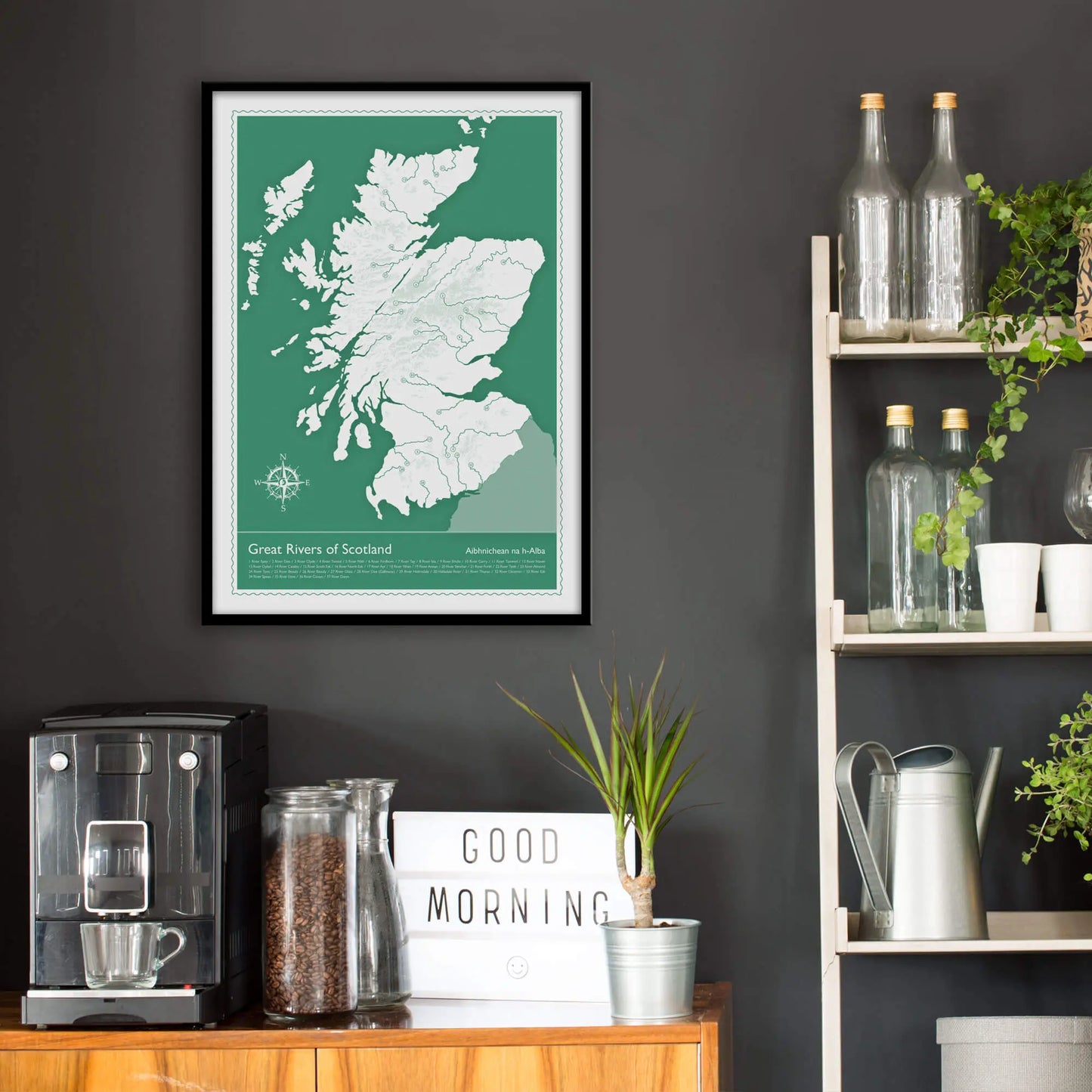 Kitchen wall infront of which is a sideboard with a coffee machine and a set of shelves holding bottles and plants. On the wall above the sideboard is the framed print in a black frame on a dark grey wall.