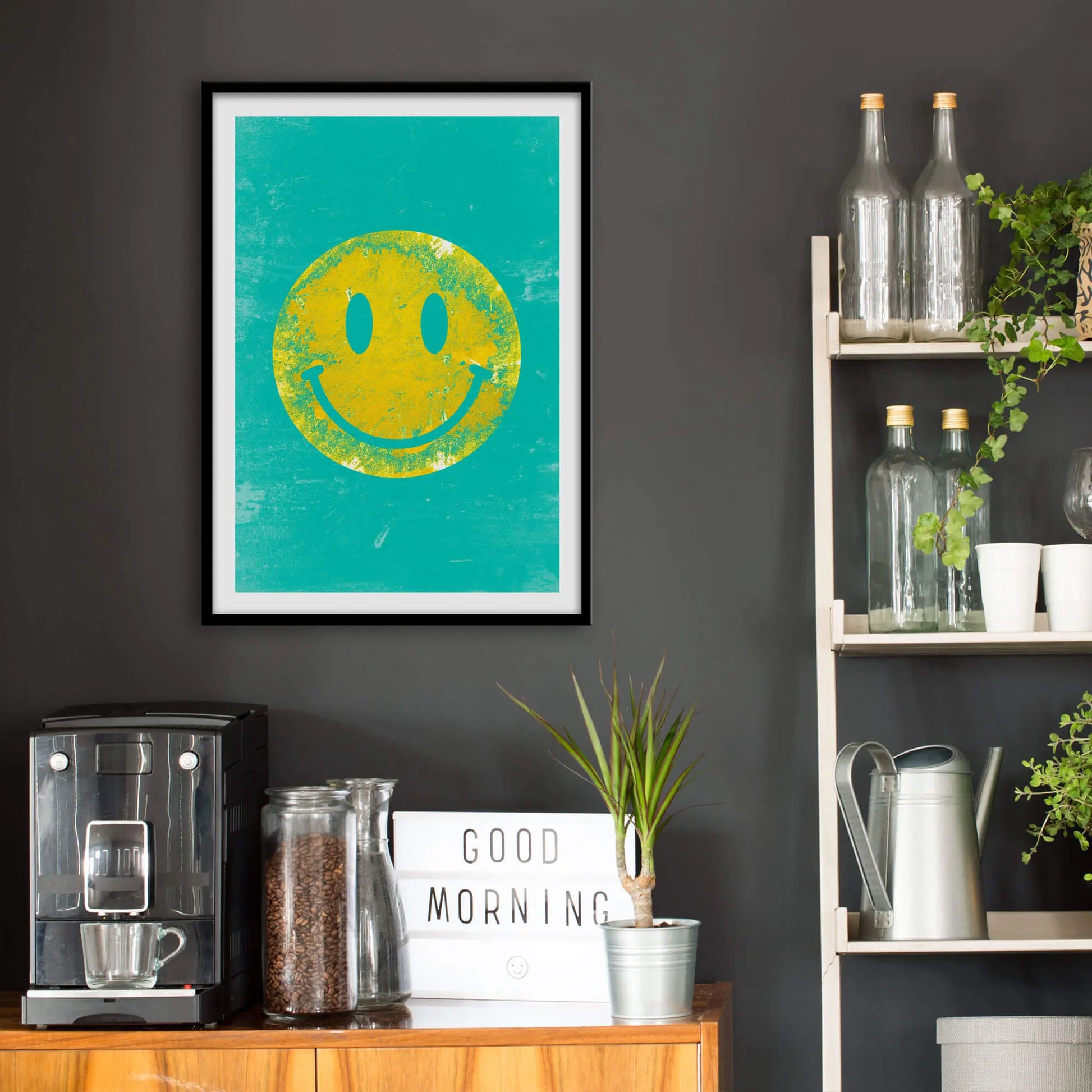 Kitchen wall infront of which is a sideboard with a coffee machine and a set of shelves holding bottles and plants. On the wall above the sideboard is the framed print in a black frame on a dark grey wall.