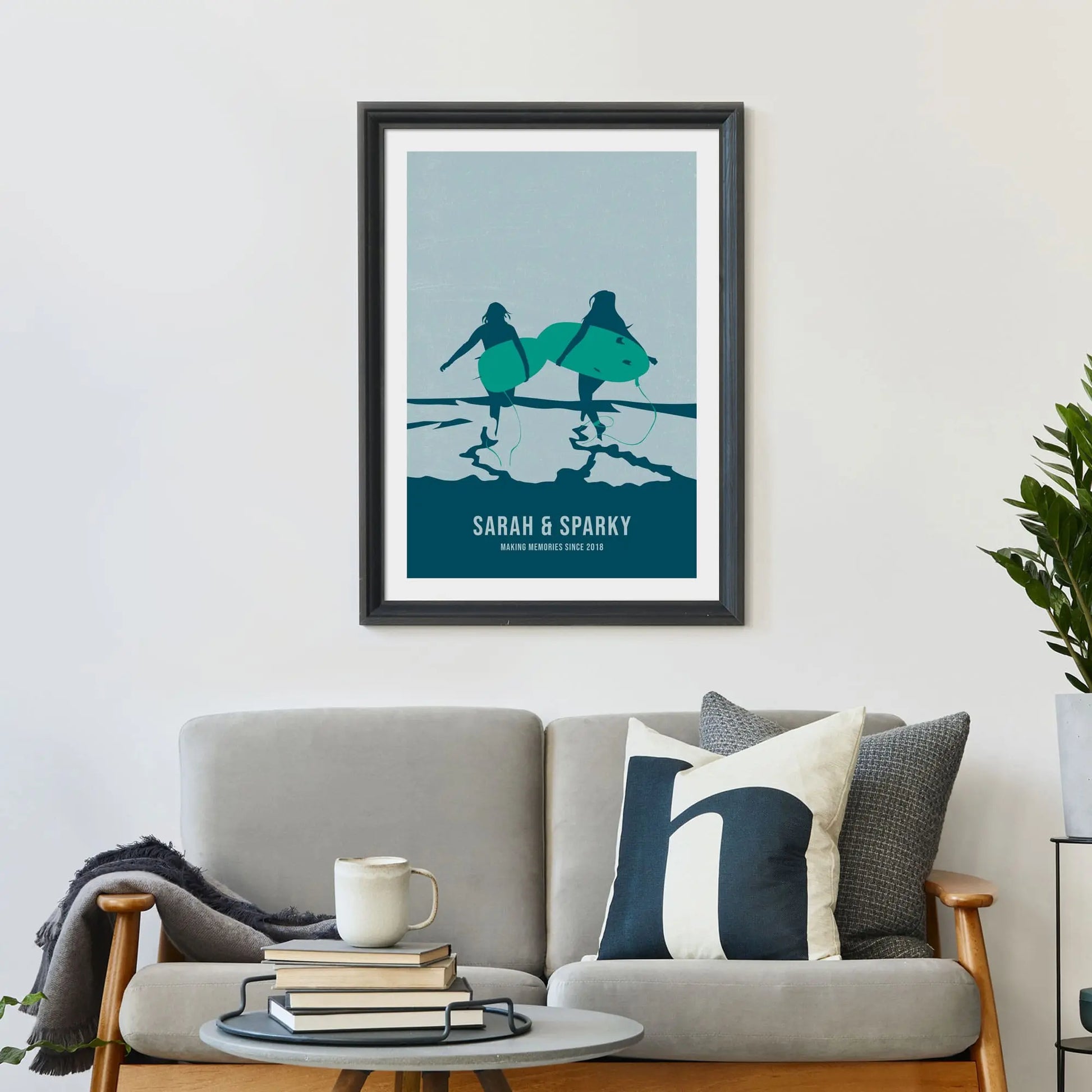 Personalised Surfing Mates Art Print in navy and green from Purest Green Prints.Large print on wall behind a 2 seater sofa covered in cushions with a coffee table covered in books.