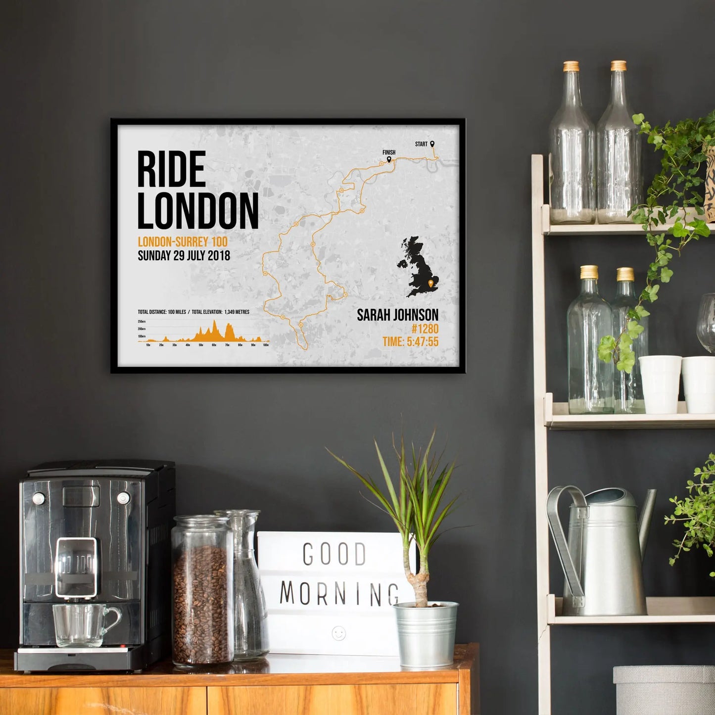 Kitchen wall infront of which is a sideboard with a coffee machine and a set of shelves holding bottles and plants. On the wall above the sideboard is the framed print in a black frame on a dark grey wall.