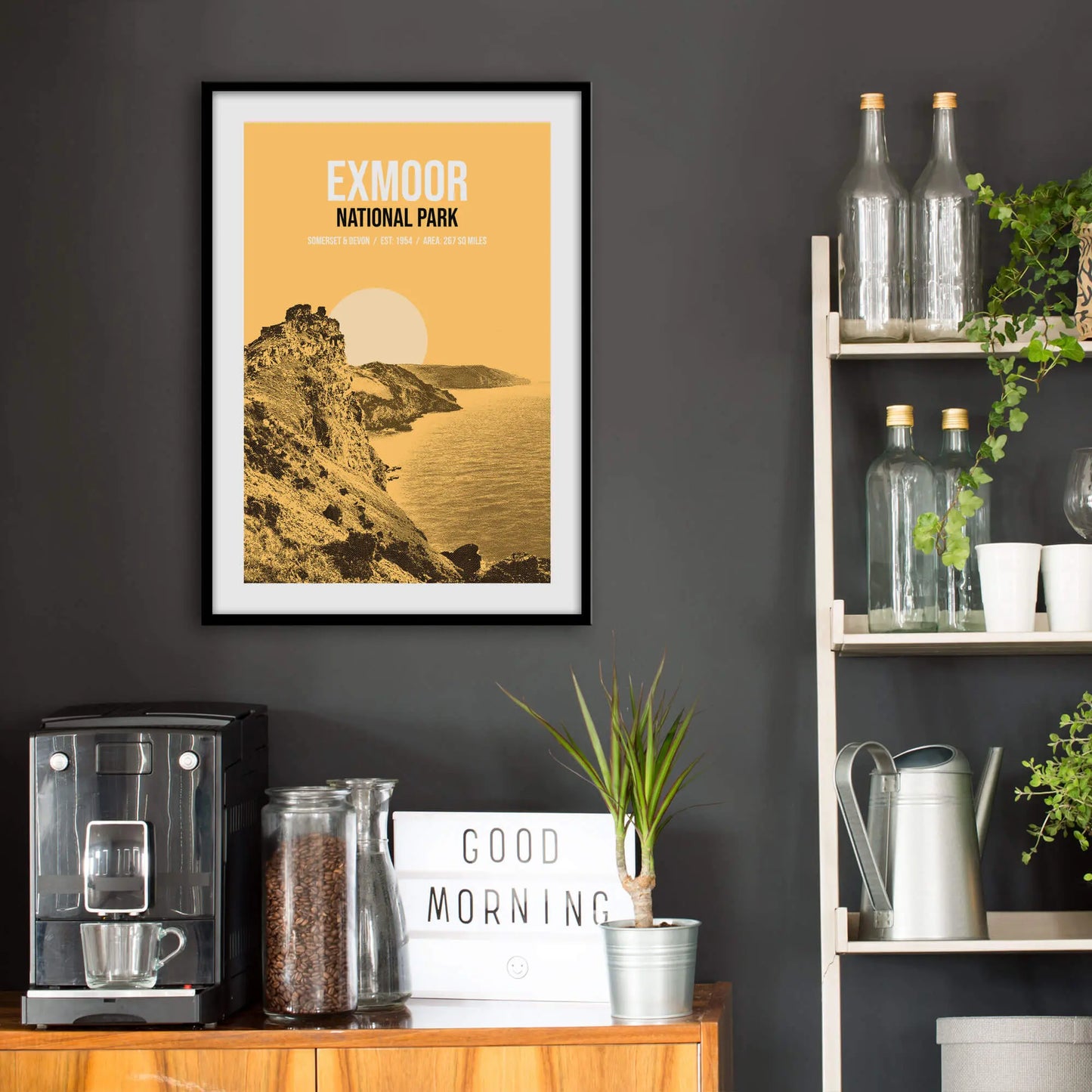 Kitchen wall infront of which is a sideboard with a coffee machine and a set of shelves holding bottles and plants. On the wall above the sideboard is the framed print in a black frame on a dark grey wall.