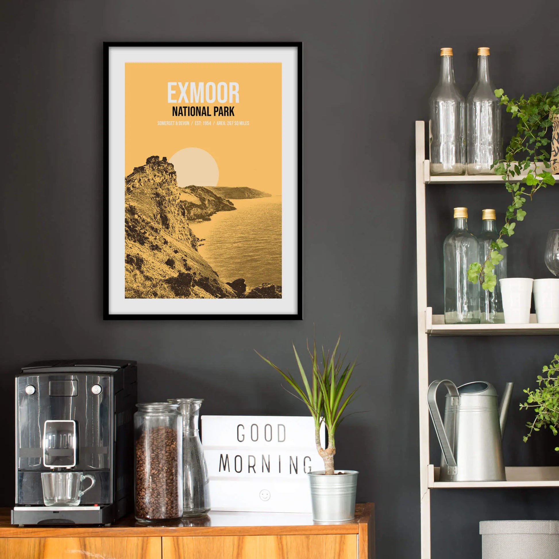 Kitchen wall infront of which is a sideboard with a coffee machine and a set of shelves holding bottles and plants. On the wall above the sideboard is the framed print in a black frame on a dark grey wall.