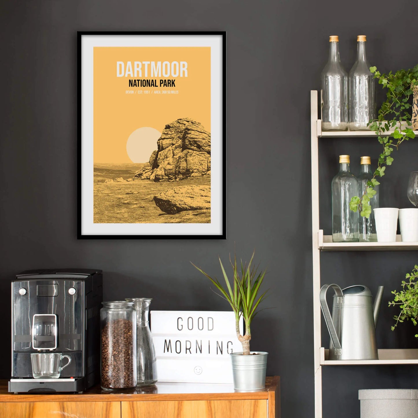 Kitchen wall infront of which is a sideboard with a coffee machine and a set of shelves holding bottles and plants. On the wall above the sideboard is the framed print in a black frame on a dark grey wall.