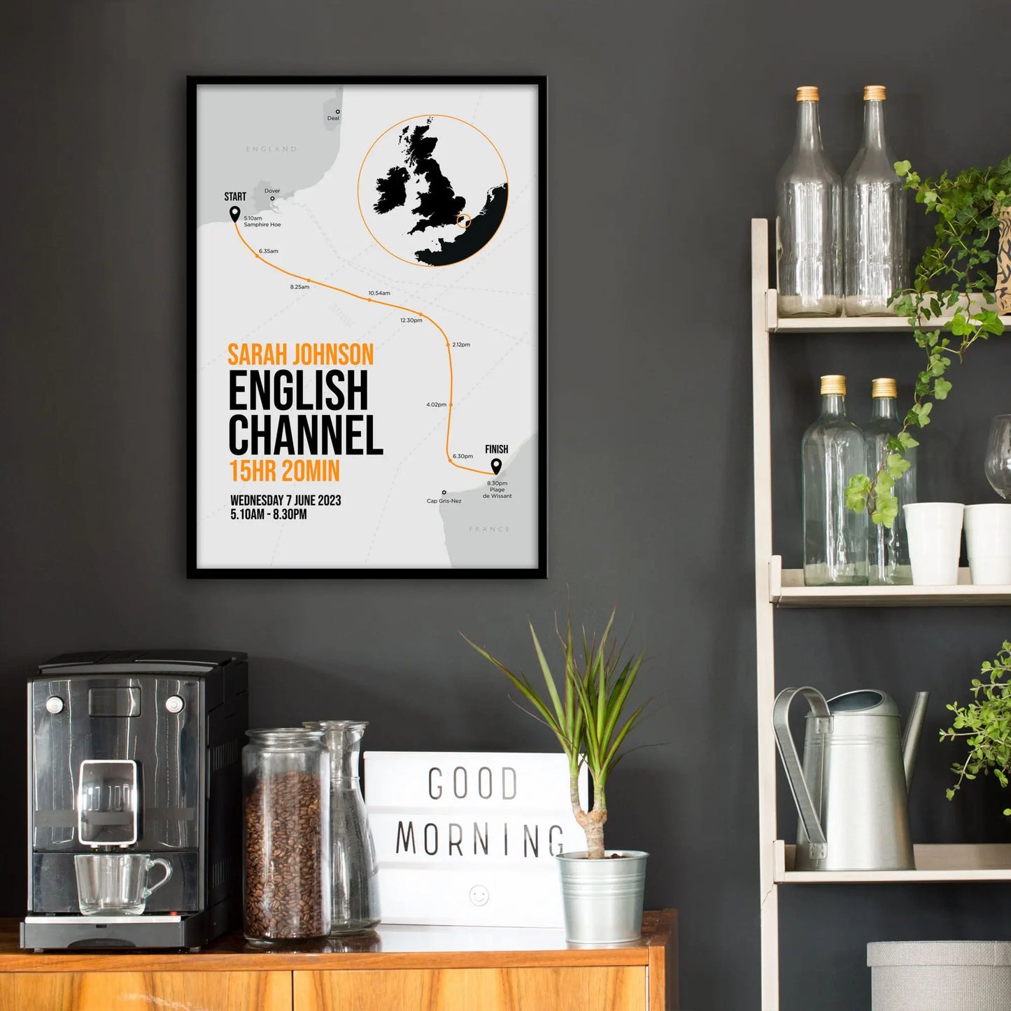 Kitchen wall infront of which is a sideboard with a coffee machine and a set of shelves holding bottles and plants. On the wall above the sideboard is the framed print in a black frame on a dark grey wall.