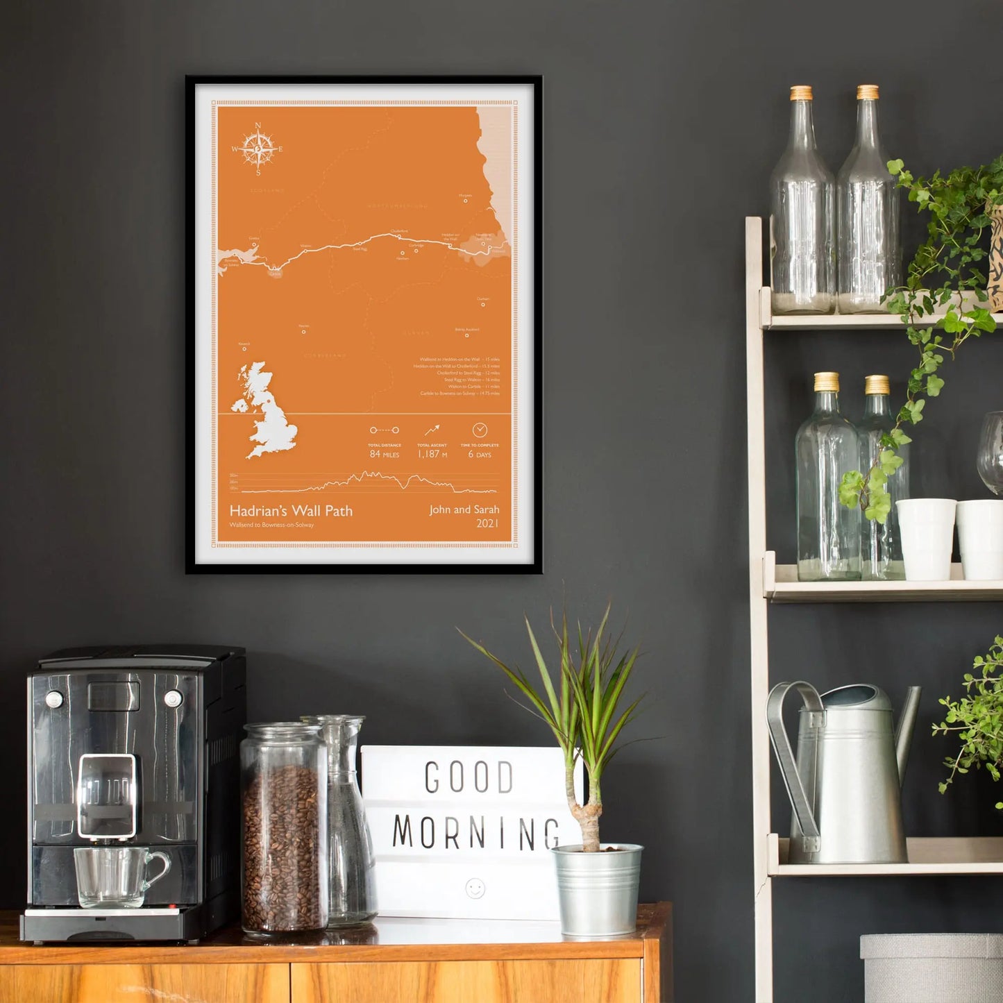 Kitchen wall infront of which is a sideboard with a coffee machine and a set of shelves holding bottles and plants. On the wall above the sideboard is the framed print in a black frame on a dark grey wall.