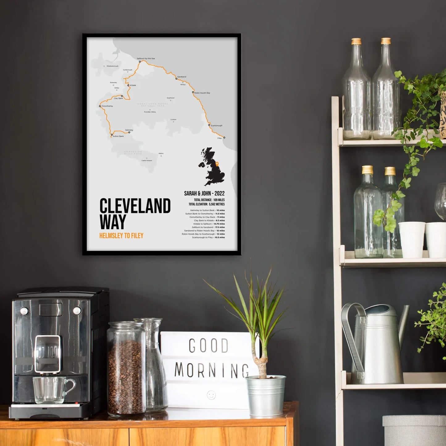 Kitchen wall infront of which is a sideboard with a coffee machine and a set of shelves holding bottles and plants. On the wall above the sideboard is the framed print in a black frame on a dark grey wall.