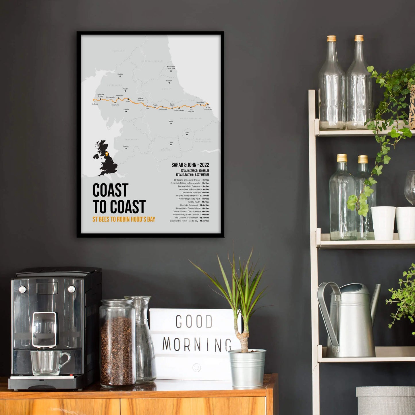 Kitchen wall infront of which is a sideboard with a coffee machine and a set of shelves holding bottles and plants. On the wall above the sideboard is the framed print in a black frame on a dark grey wall.