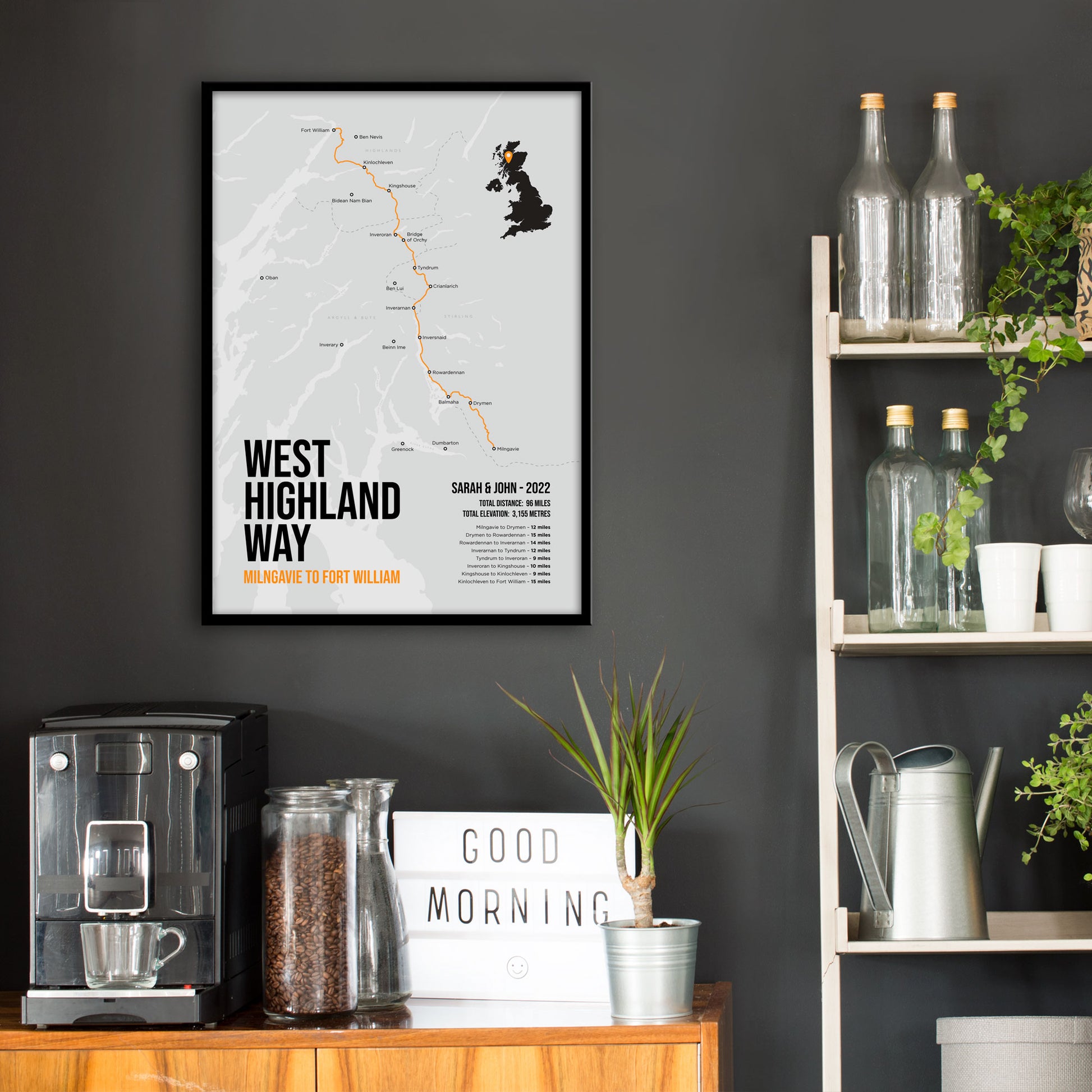 Kitchen wall infront of which is a sideboard with a coffee machine and a set of shelves holding bottles and plants. On the wall above the sideboard is the framed print in a black frame on a dark grey wall.