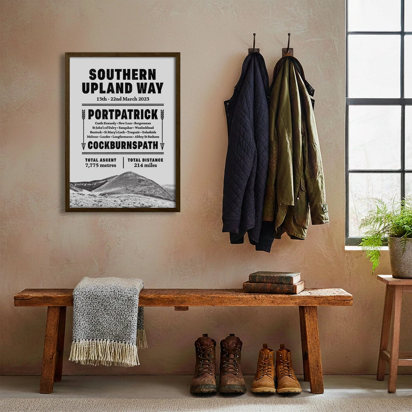 Framed print hanging on cream wall beside coats hanging above bench with walking boots and a scarf