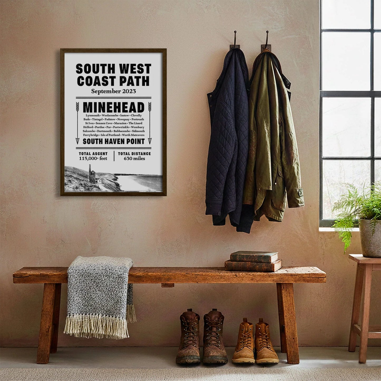 Framed print hanging on cream wall beside coats hanging above bench with walking boots and a scarf