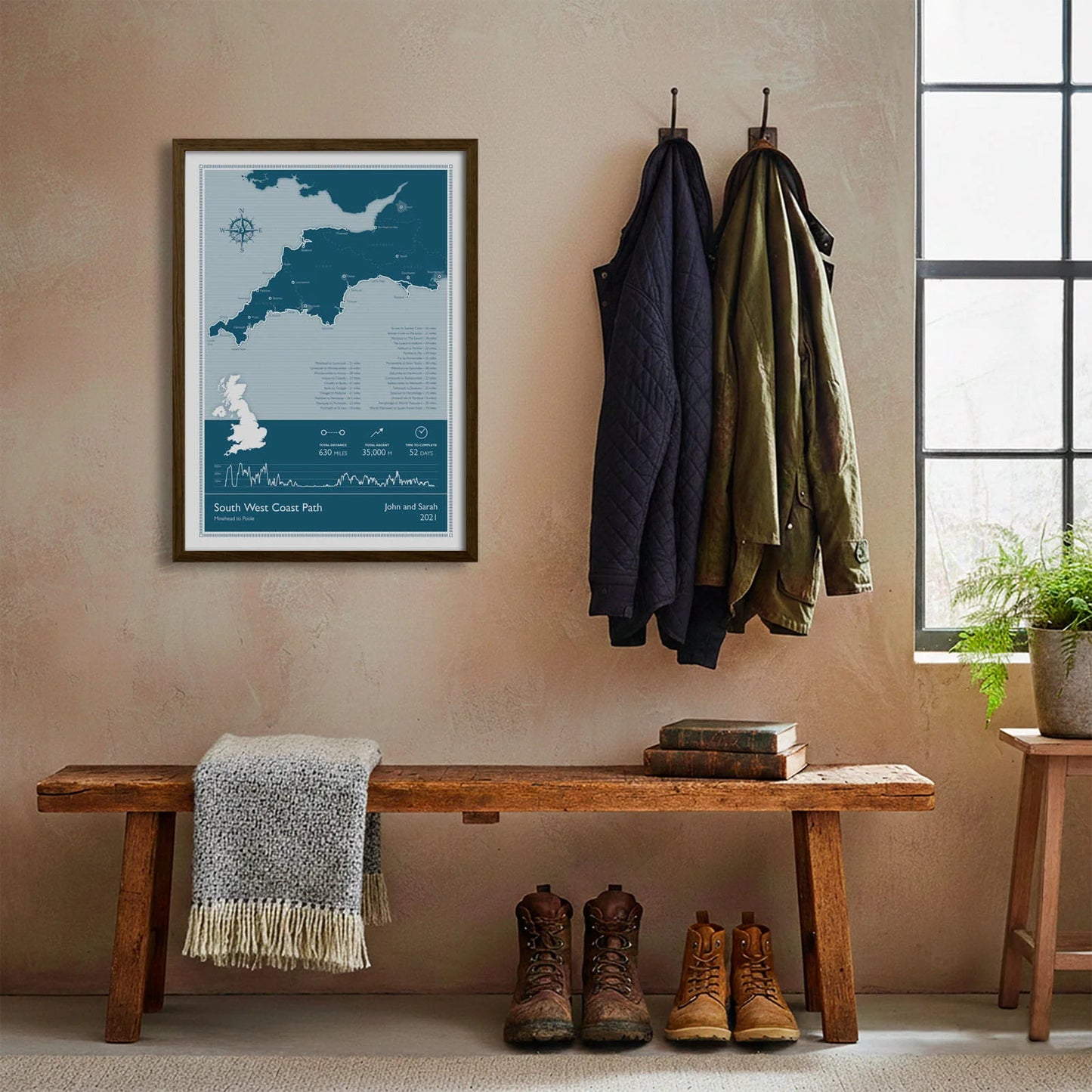 Framed print hanging on cream wall beside coats hanging above bench with walking boots and a scarf