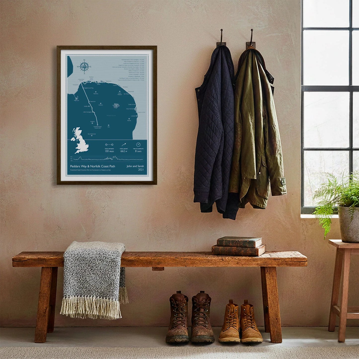 Framed print hanging on cream wall beside coats hanging above bench with walking boots and a scarf