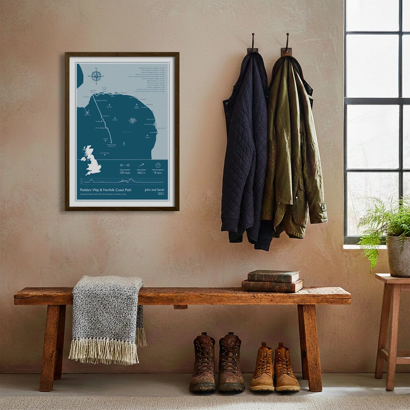 Framed print hanging on cream wall beside coats hanging above bench with walking boots and a scarf