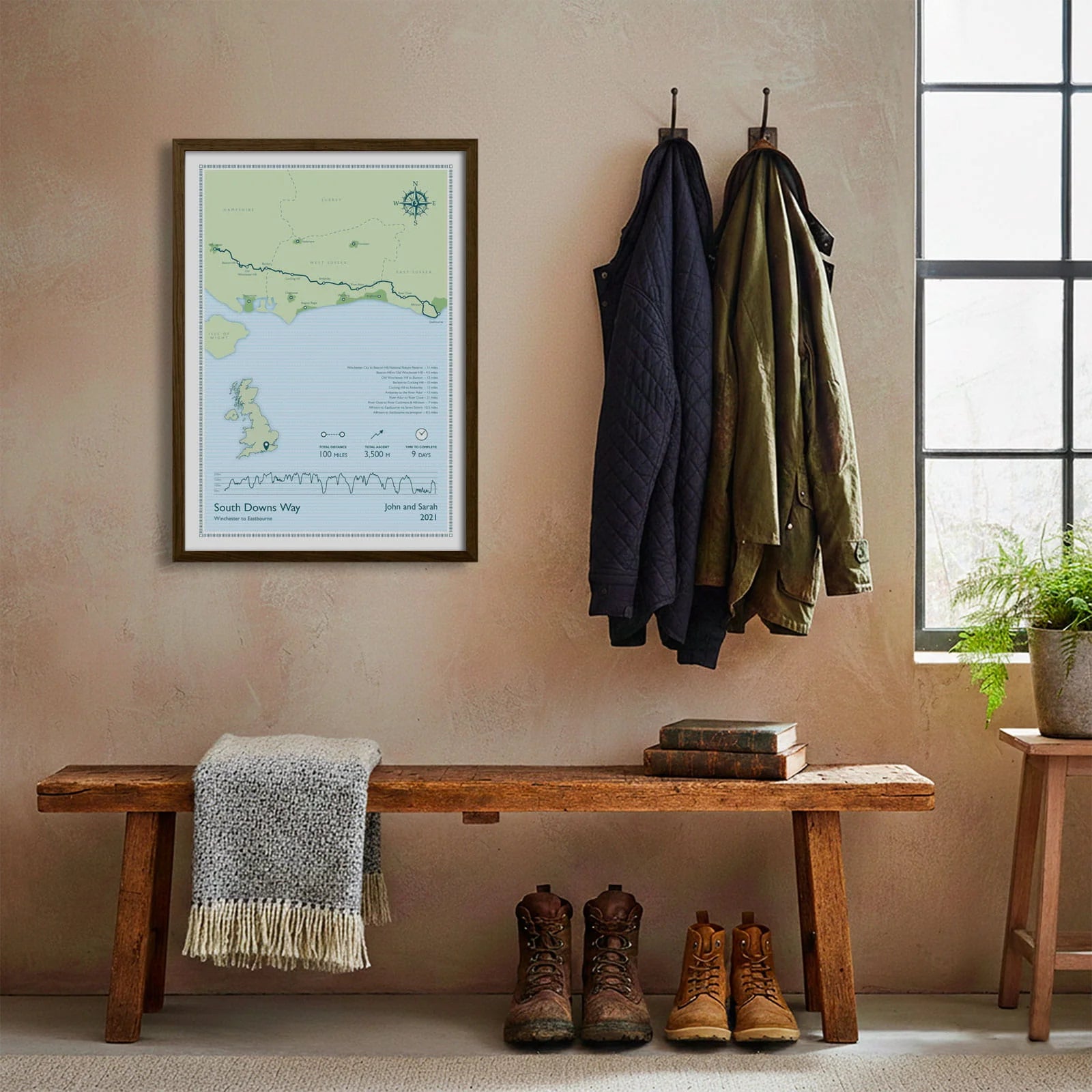 Framed print hanging on cream wall beside coats hanging above bench with walking boots and a scarf