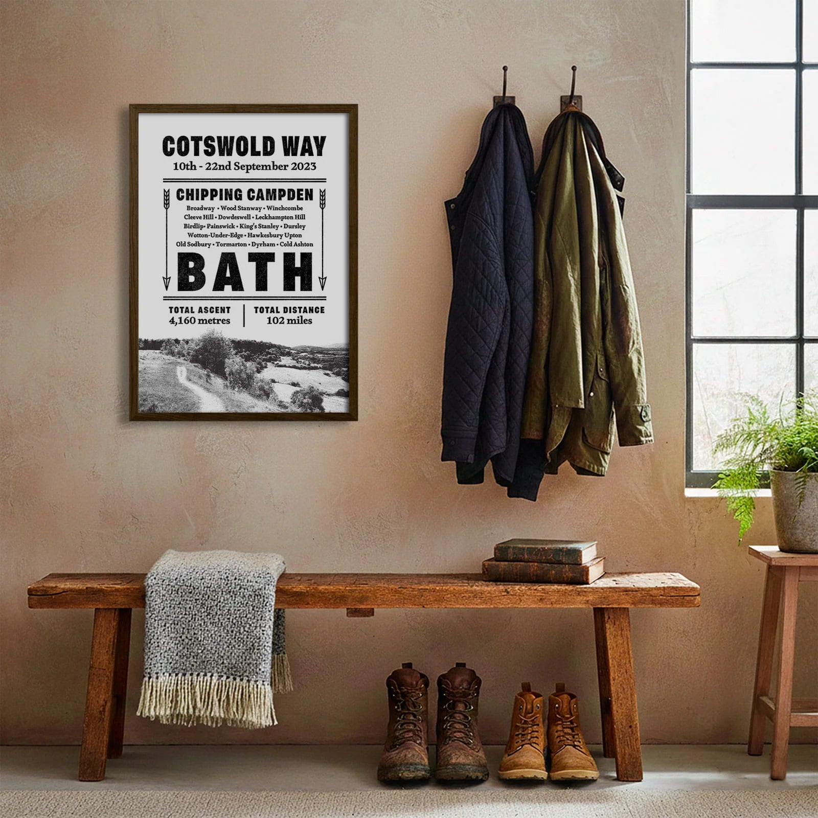 Framed print hanging on cream wall beside coats hanging above bench with walking boots and a scarf