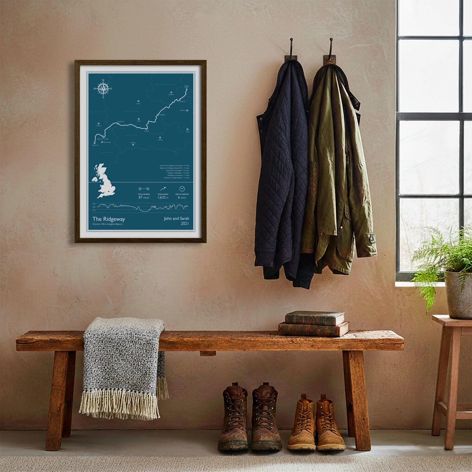 Framed print hanging on cream wall beside coats hanging above bench with walking boots and a scarf