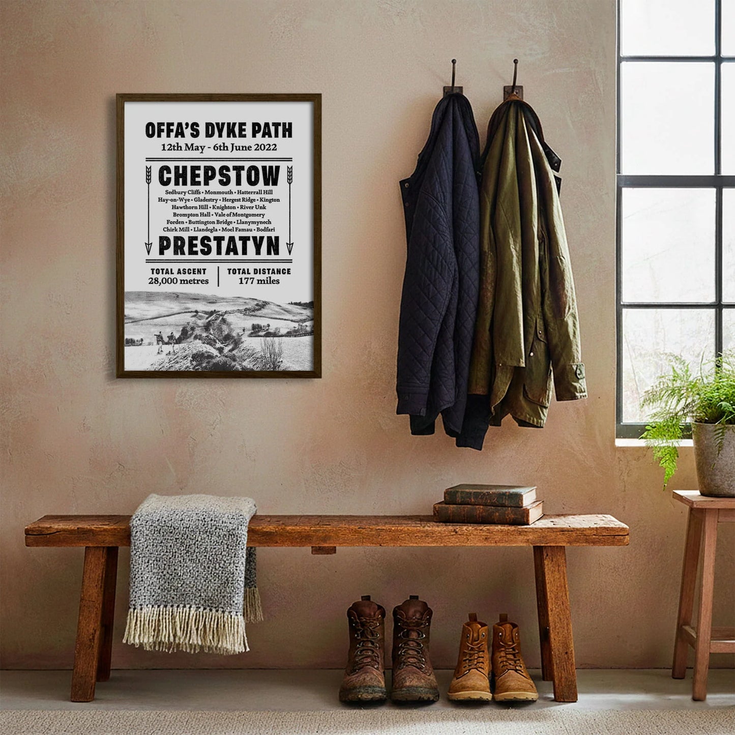 Framed print hanging on cream wall beside coats hanging above bench with walking boots and a scarf