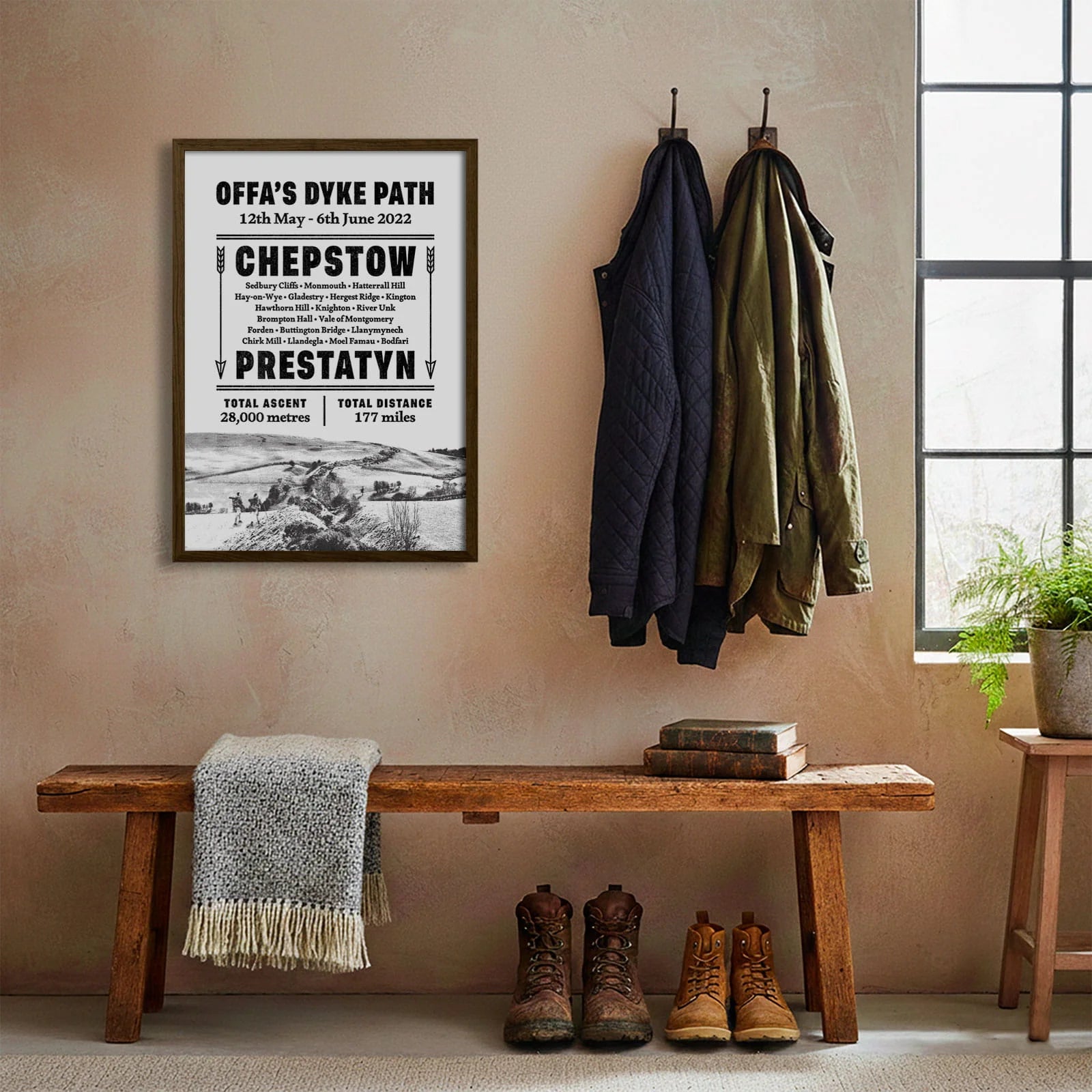 Framed print hanging on cream wall beside coats hanging above bench with walking boots and a scarf
