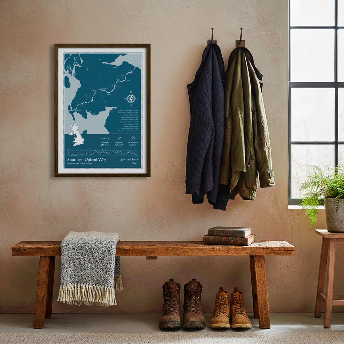 Framed print hanging on cream wall beside coats hanging above bench with walking boots and a scarf