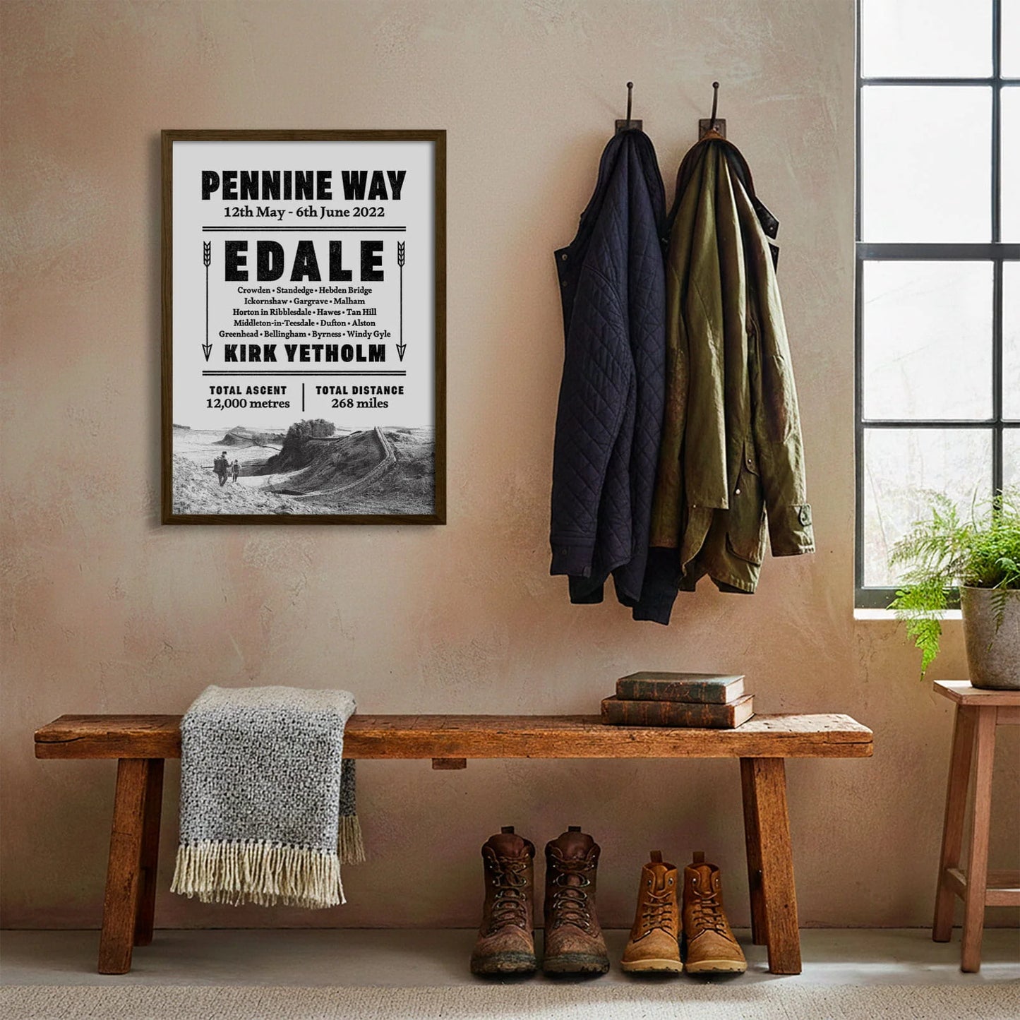 Framed print hanging on cream wall beside coats hanging above bench with walking boots and a scarf