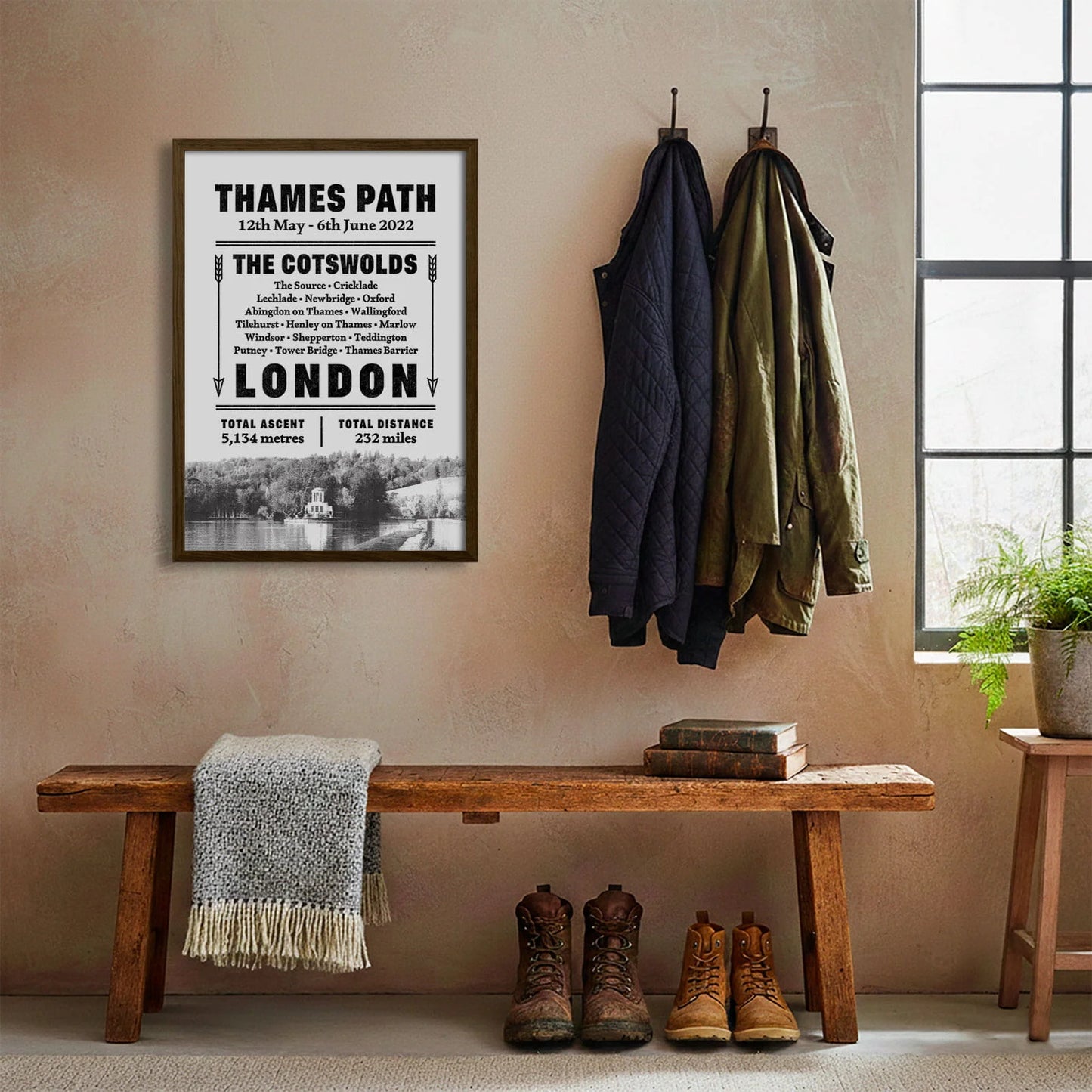 Framed print hanging on cream wall beside coats hanging above bench with walking boots and a scarf