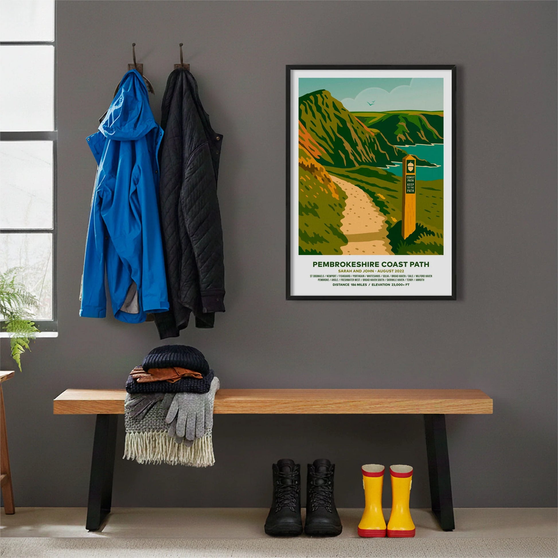 Framed print hanging on grey wall beside coats hanging above bench with walking boots and a scarf