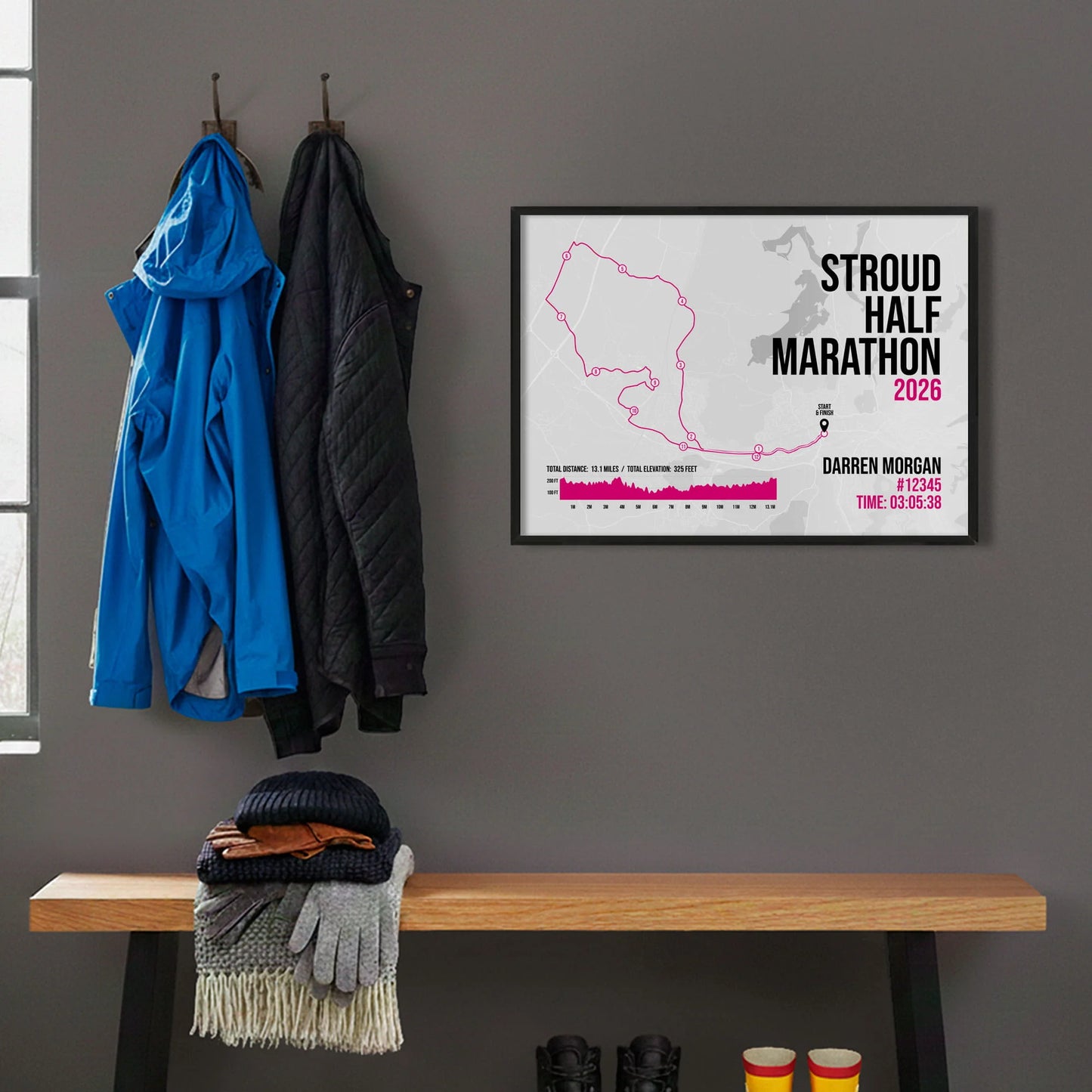Framed print on grey wall beside hanging coats above a bench with walking boots