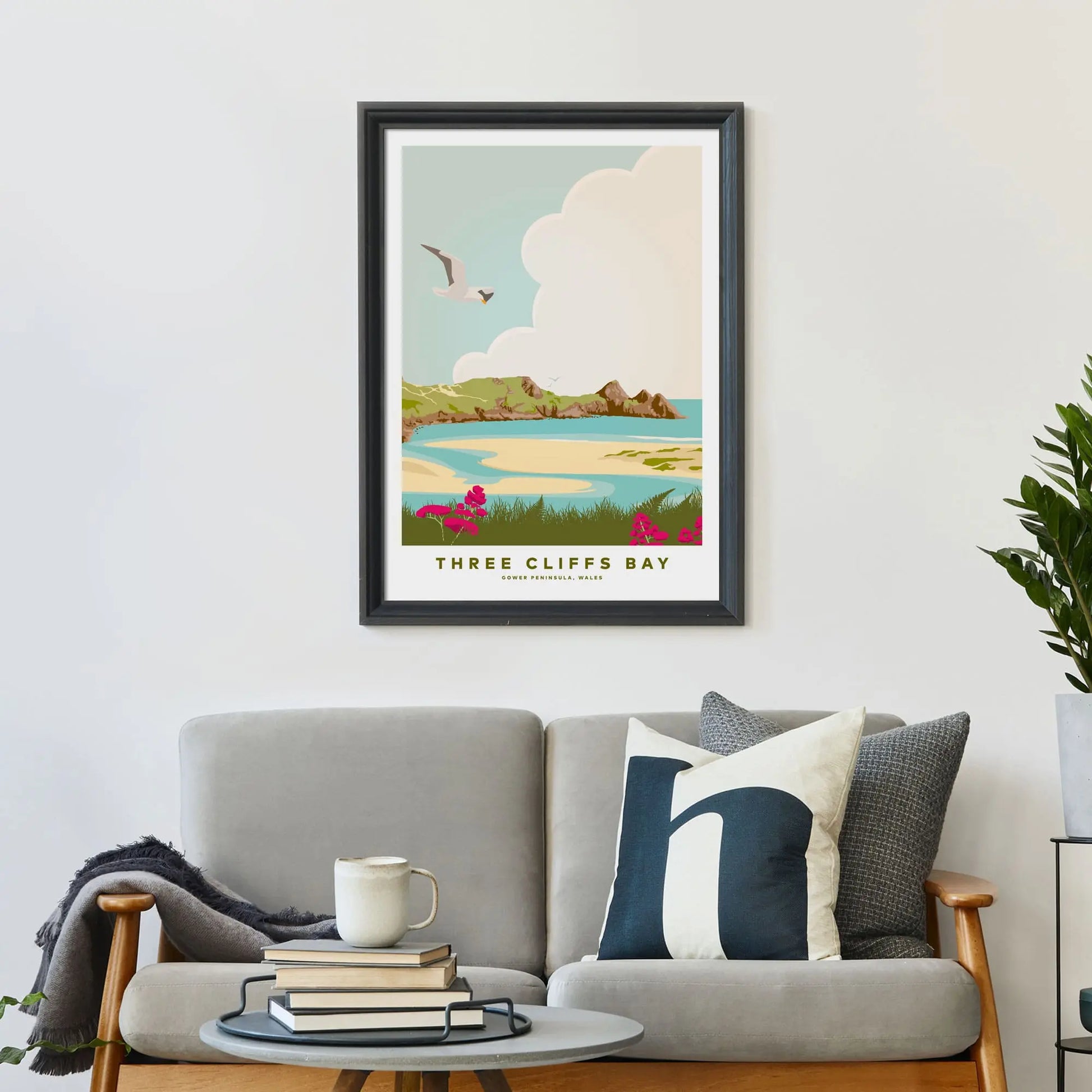 Three Cliffs print framed on wall above a sofa covered in cushions with a coffee table in front.