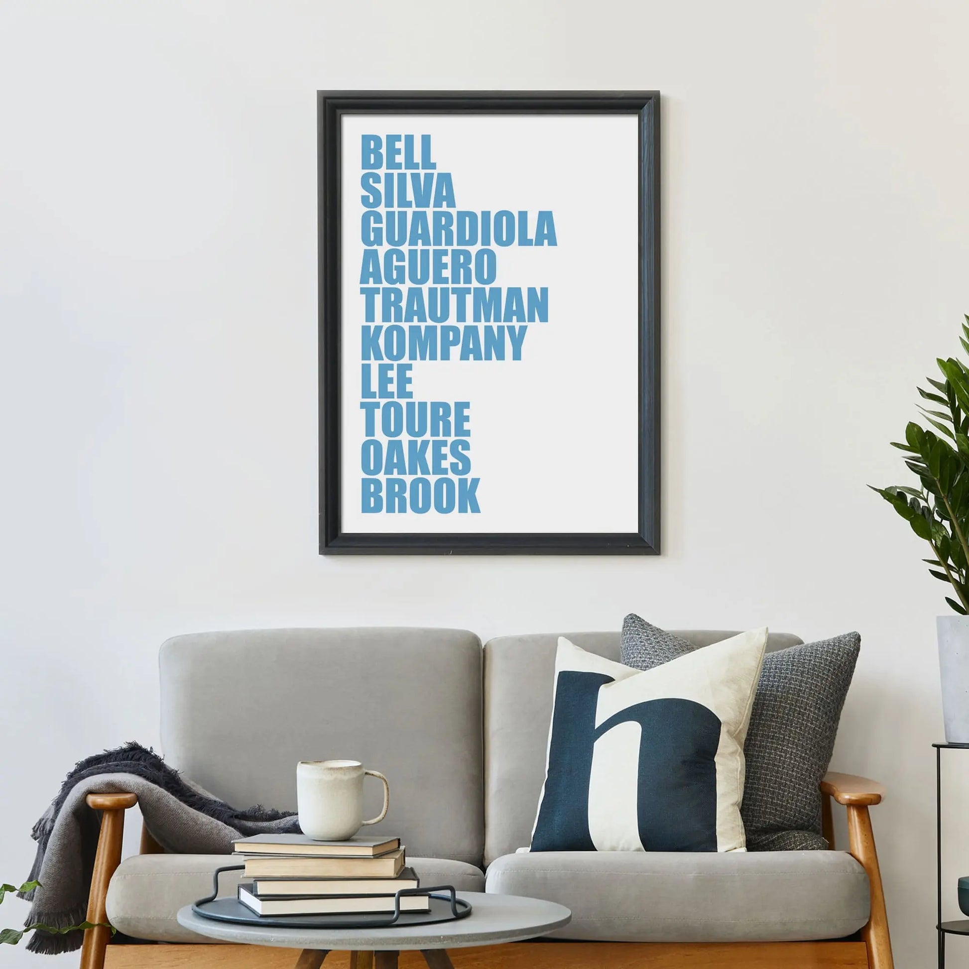 Large print on wall behind a 2 seater sofa covered in cushions with a coffee table covered in books.