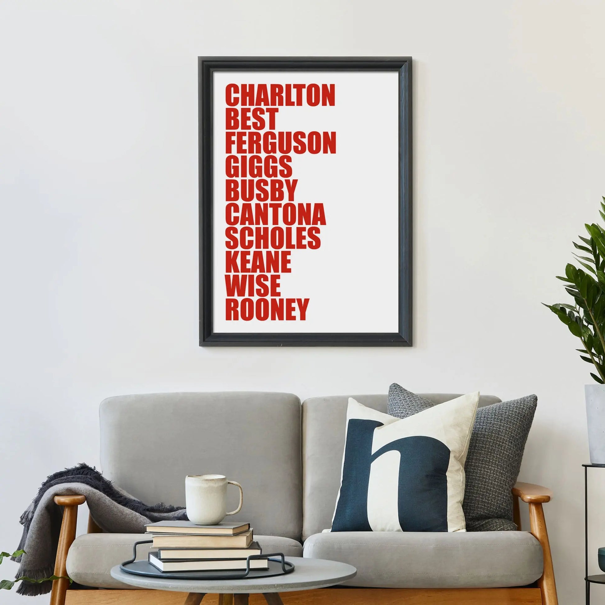 Large print on wall behind a 2 seater sofa covered in cushions with a coffee table covered in books.