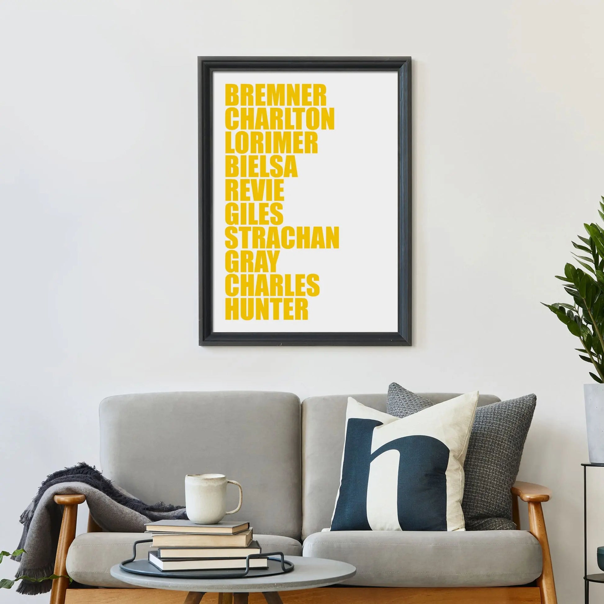 Large print on wall behind a 2 seater sofa covered in cushions with a coffee table covered in books.