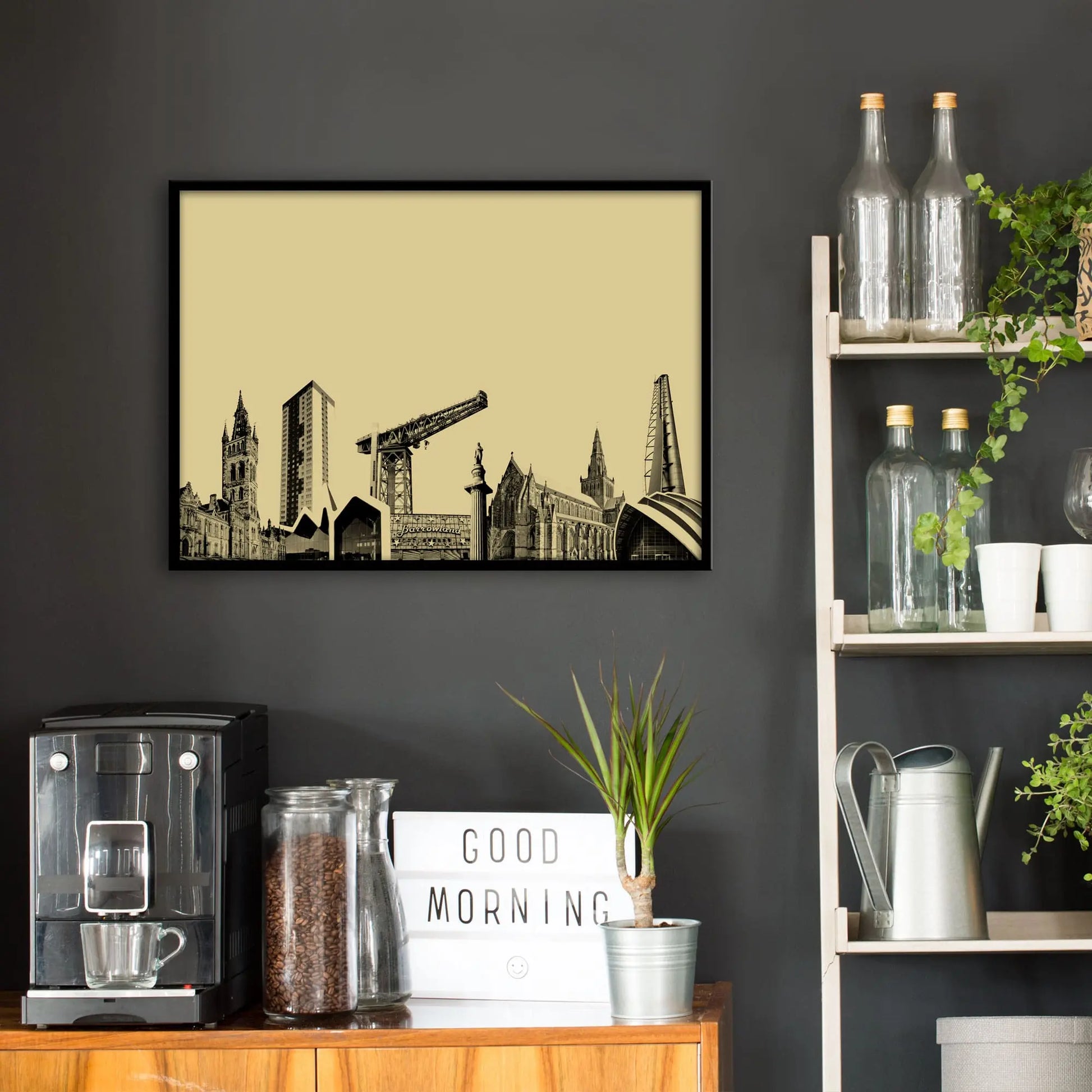 Kitchen wall infront of which is a sideboard with a coffee machine and a set of shelves holding bottles and plants. On the wall above the sideboard is the framed print in a black frame on a dark grey wall.