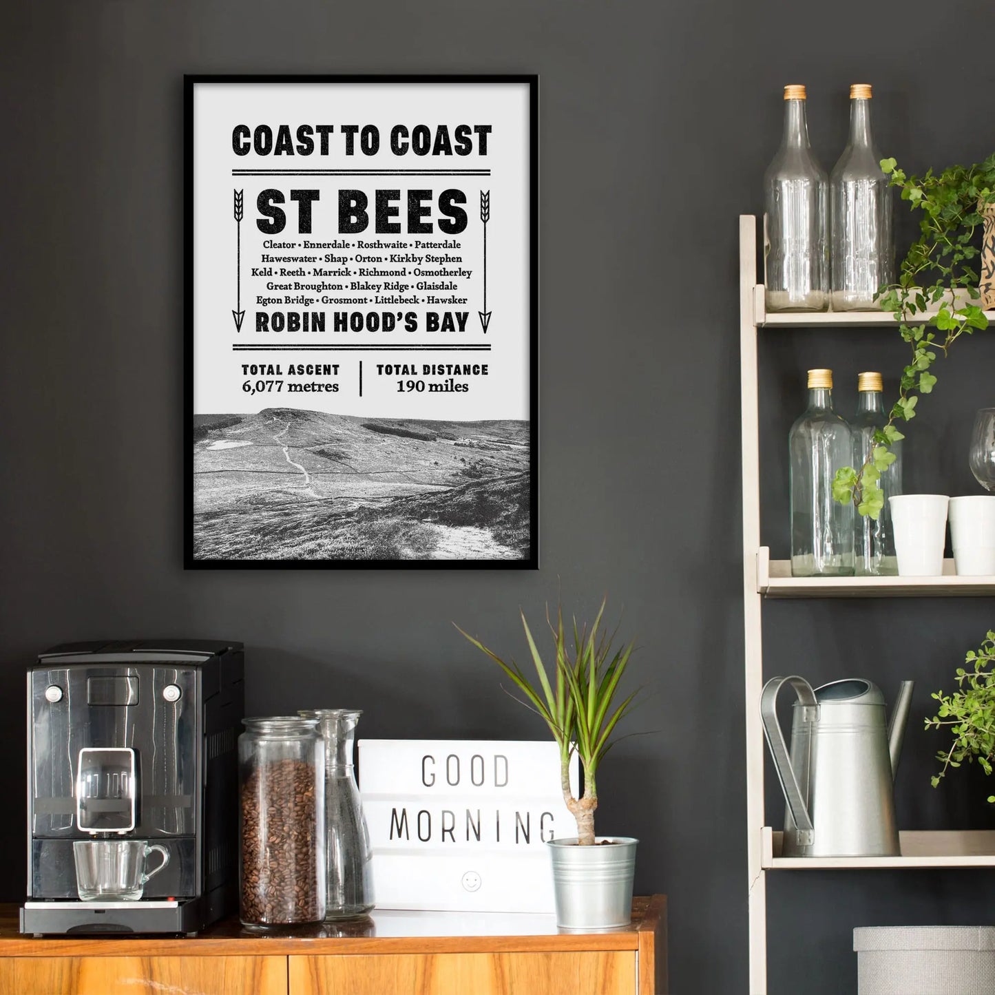 Kitchen wall infront of which is a sideboard with a coffee machine and a set of shelves holding bottles and plants. On the wall above the sideboard is the framed print in a black frame on a dark grey wall.