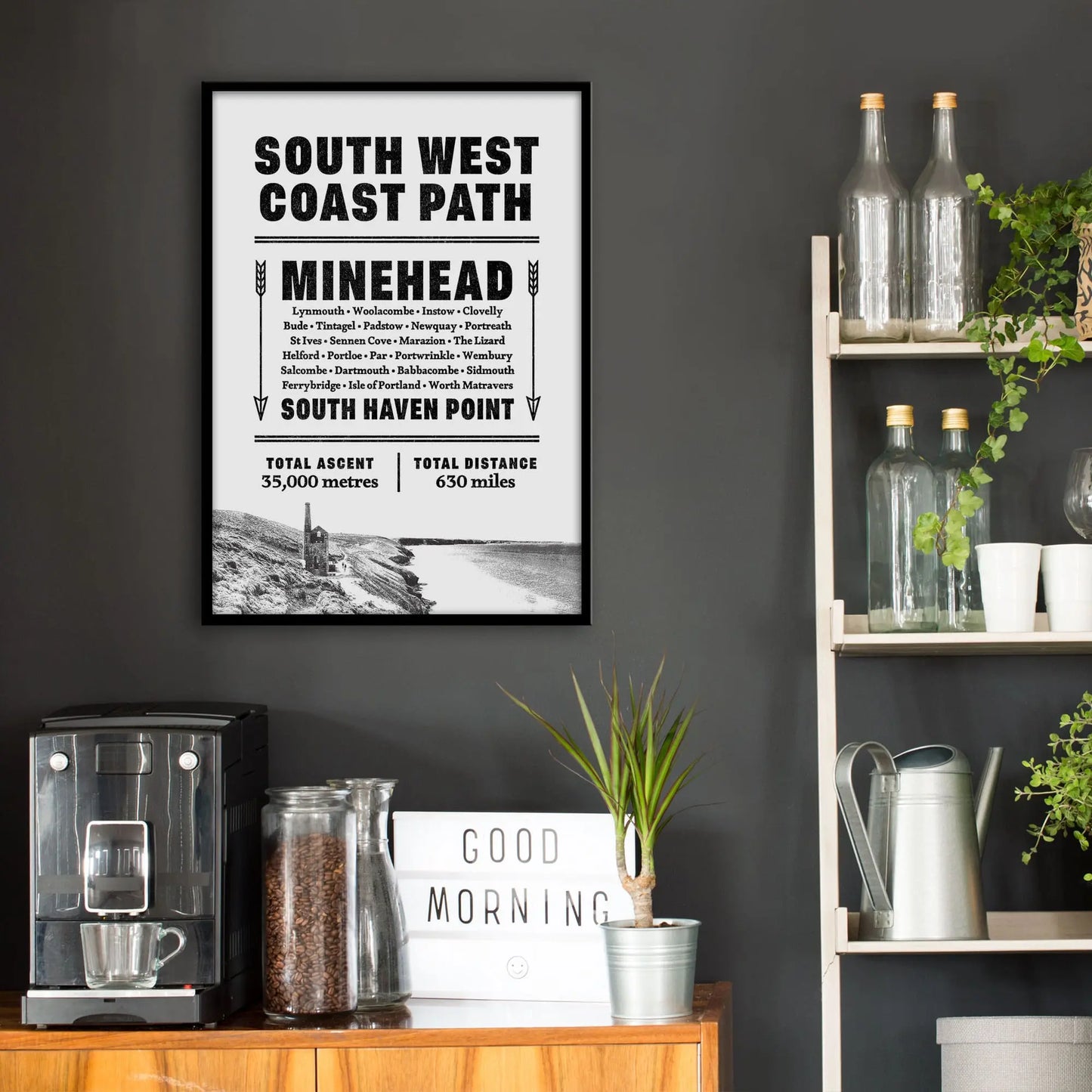 Kitchen wall infront of which is a sideboard with a coffee machine and a set of shelves holding bottles and plants. On the wall above the sideboard is the framed print in a black frame on a dark grey wall.