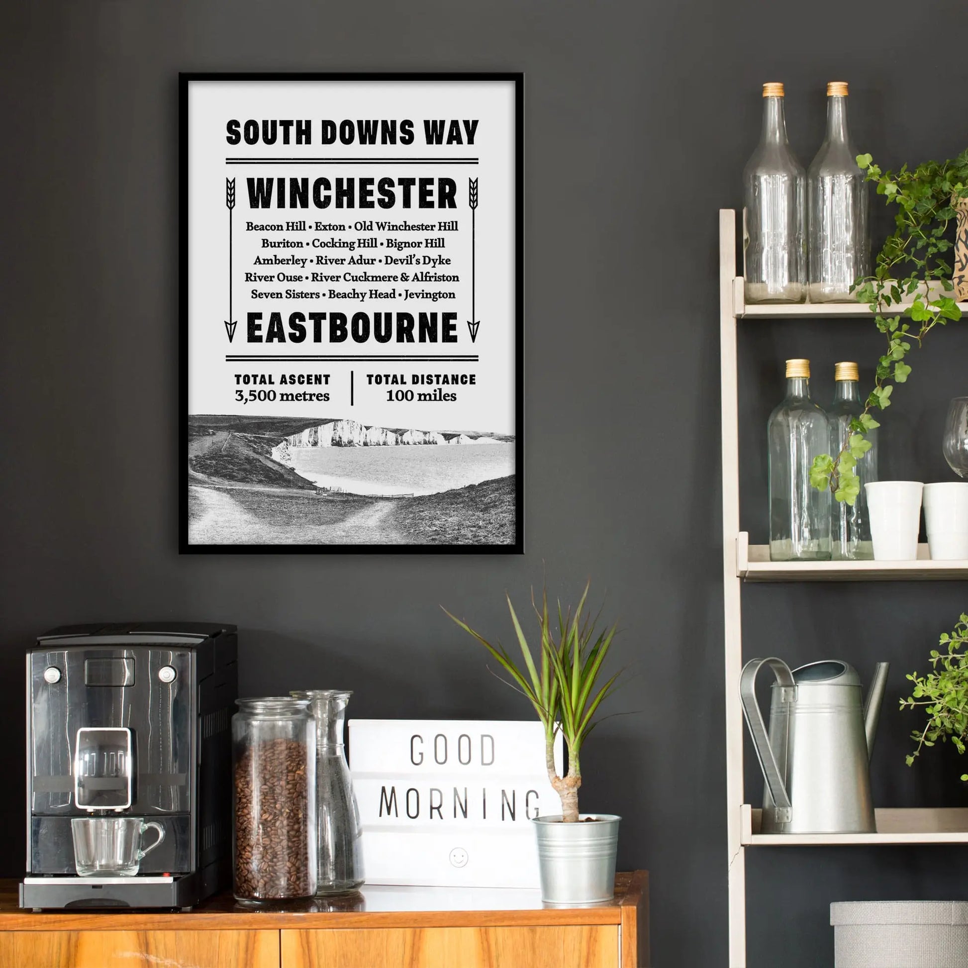 Kitchen wall infront of which is a sideboard with a coffee machine and a set of shelves holding bottles and plants. On the wall above the sideboard is the framed print in a black frame on a dark grey wall.