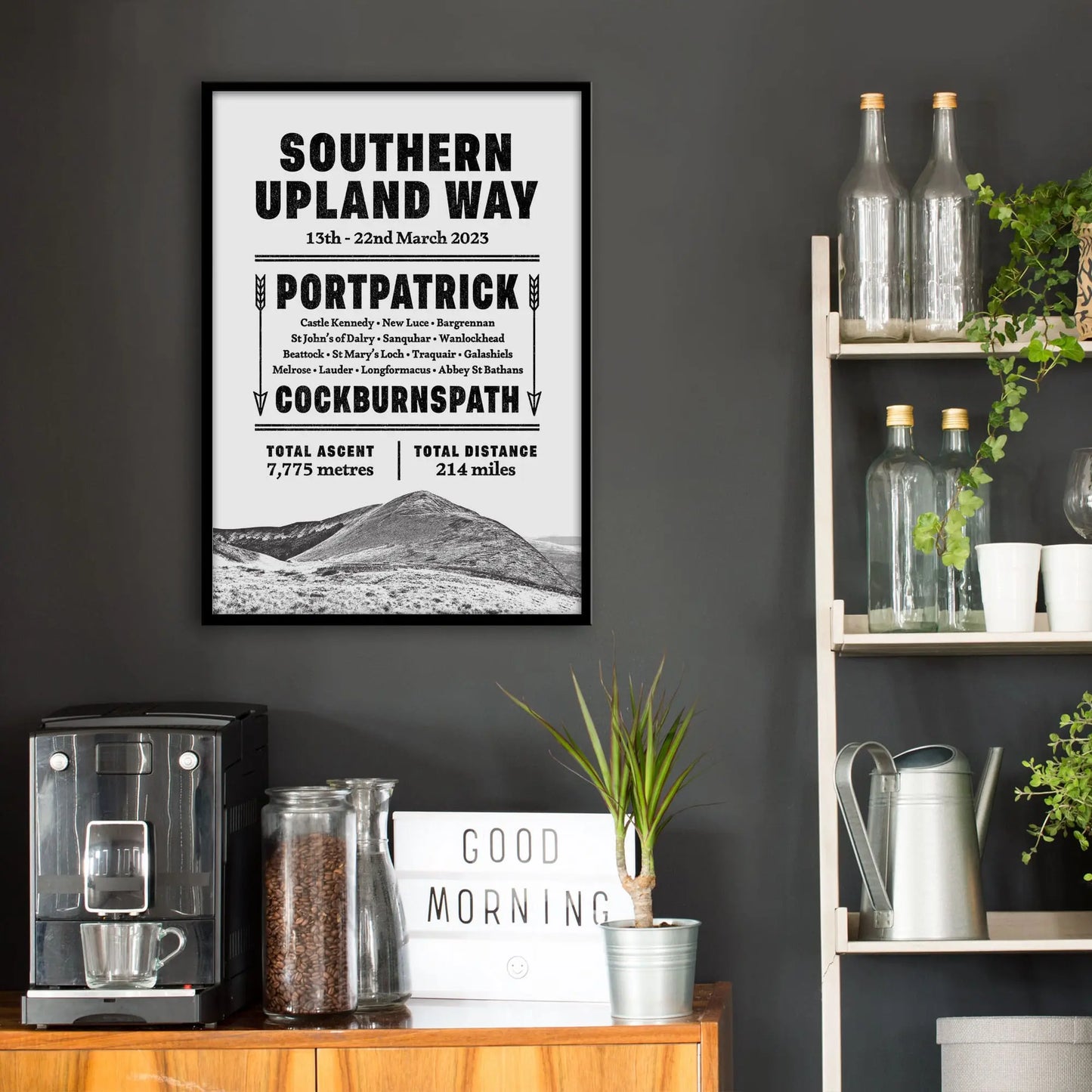 Kitchen wall infront of which is a sideboard with a coffee machine and a set of shelves holding bottles and plants. On the wall above the sideboard is the framed print in a black frame on a dark grey wall.