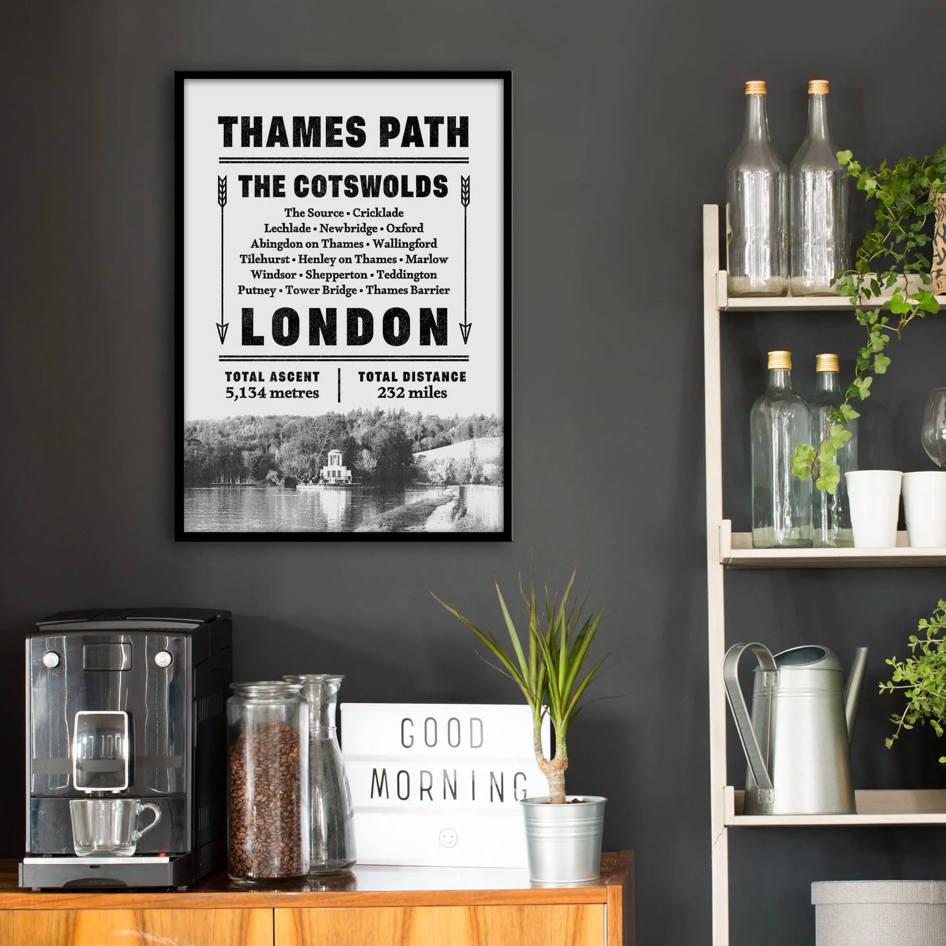 Kitchen wall infront of which is a sideboard with a coffee machine and a set of shelves holding bottles and plants. On the wall above the sideboard is the framed print in a black frame on a dark grey wall.