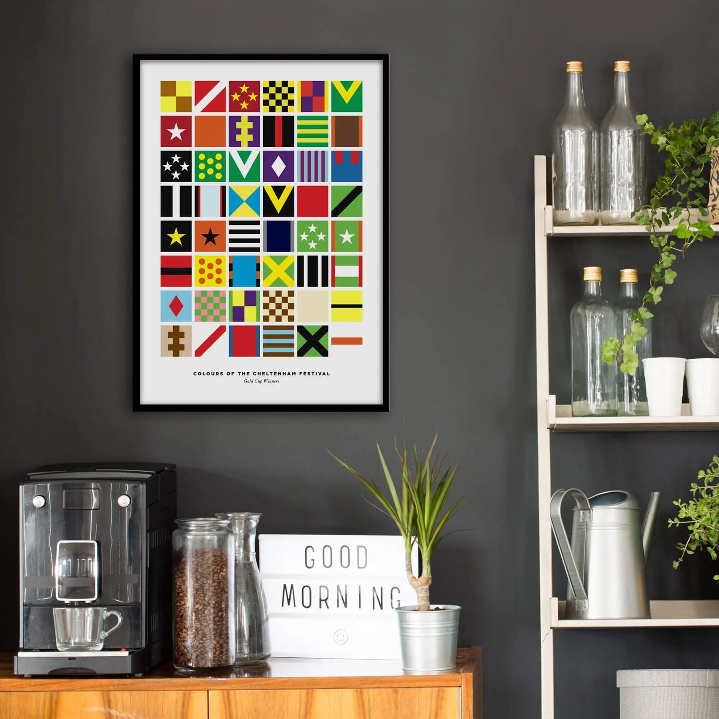 Kitchen wall infront of which is a sideboard with a coffee machine and a set of shelves holding bottles and plants. On the wall above the sideboard is the framed print in a black frame on a dark grey wall.